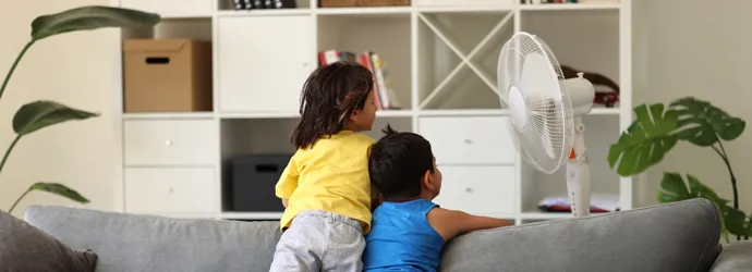 Due bambini si rinfrescano con un ventilatore affacciati allo schienale di un divano grigio.