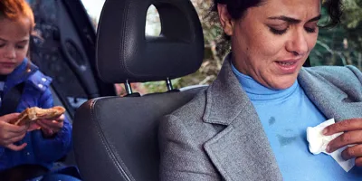 Eine Frau sitzt auf dem Vordersitz eines Autos und reinigt ihre Bluse; ein Mädchen isst ein Sandwich auf dem Rücksitz.
