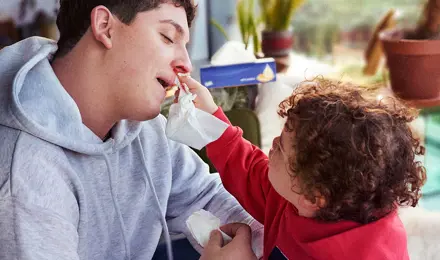 Ein kleines Kind in einer roten Latzhose hilft einem jungen Mann in einer Latzhose, sich die blutige Nase zu putzen.