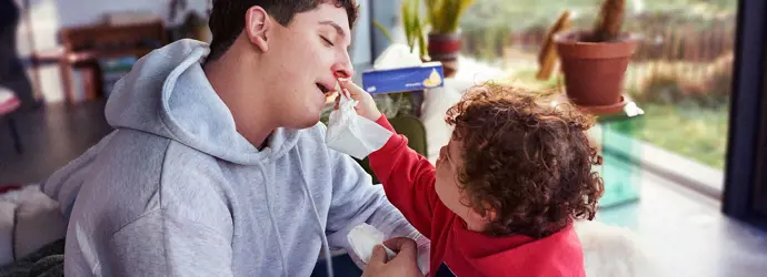 Ein kleines Kind in einer roten Latzhose hilft einem jungen Mann in einer Latzhose, sich die blutige Nase zu putzen.