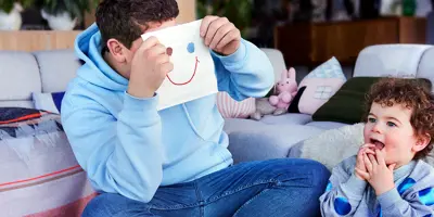 Uomo è seduto con un bambino sul pavimento e tiene in mano un fazzoletto con un volto sorridente disegnato sul davanti.