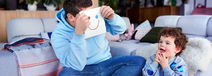 Uomo è seduto con un bambino sul pavimento e tiene in mano un fazzoletto con un volto sorridente disegnato sul davanti.
