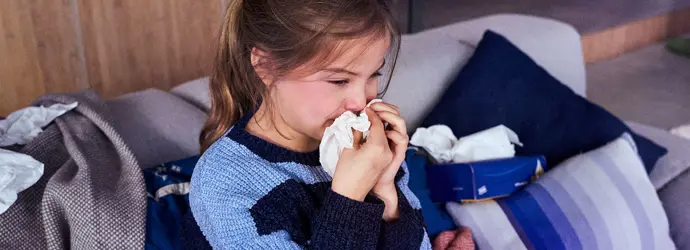 Una ragazza siede su un divano ed è circondata da fazzoletti mentre starnutisce in un fazzoletto.